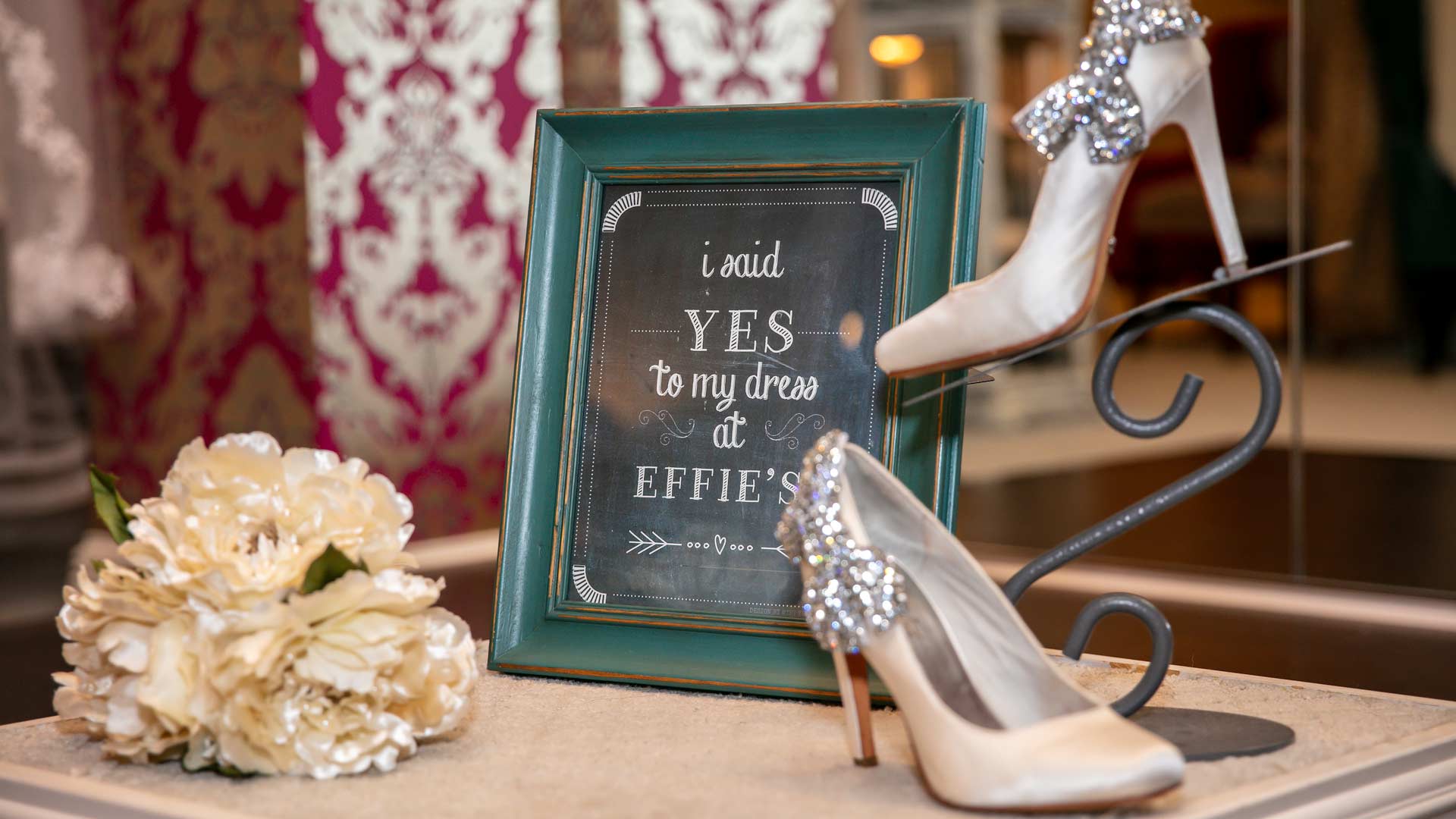 Bridal shoes and a bouquet on a table with chalkboard with "I said YES to my dress at Effie's"